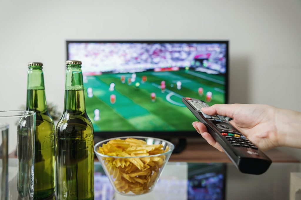 A man using a TV remote to switch on Fox Sports to watch a football match. 